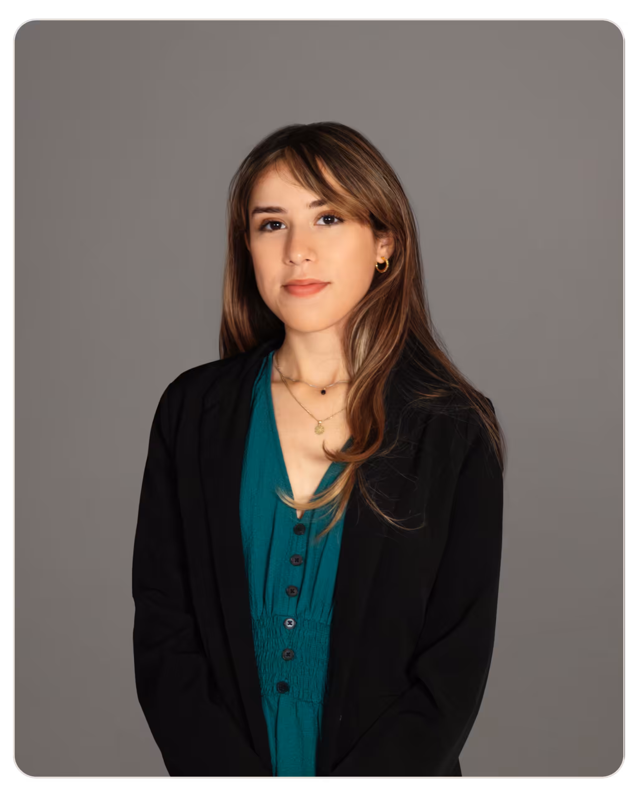 Kandela Adela Ramos MSW. Portrait of a woman with light brown hair wearing a teal blouse and black blazer against a gray studio backdrop, wearing gold hoop earrings and layered necklaces.