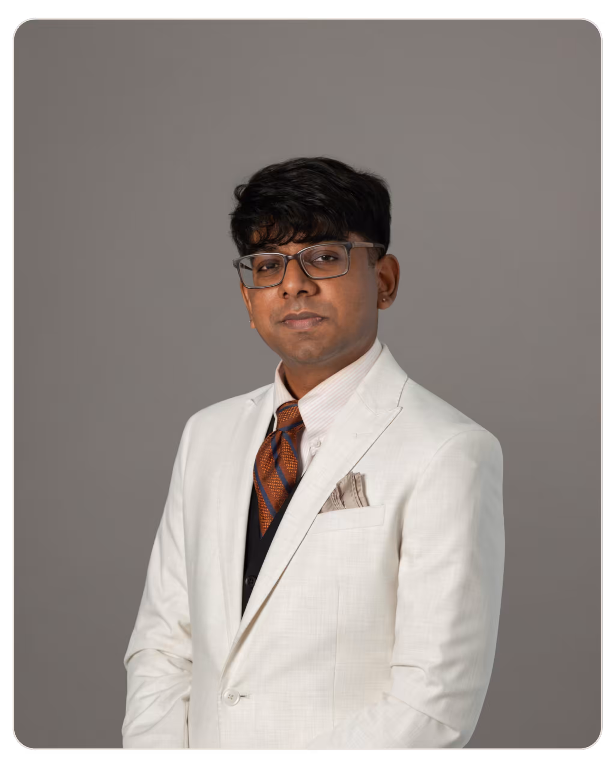 Aldrin DSilva, MA Portrait of a man in a cream suit, striped brown-orange tie, and glasses, posing against a gray studio backdrop.