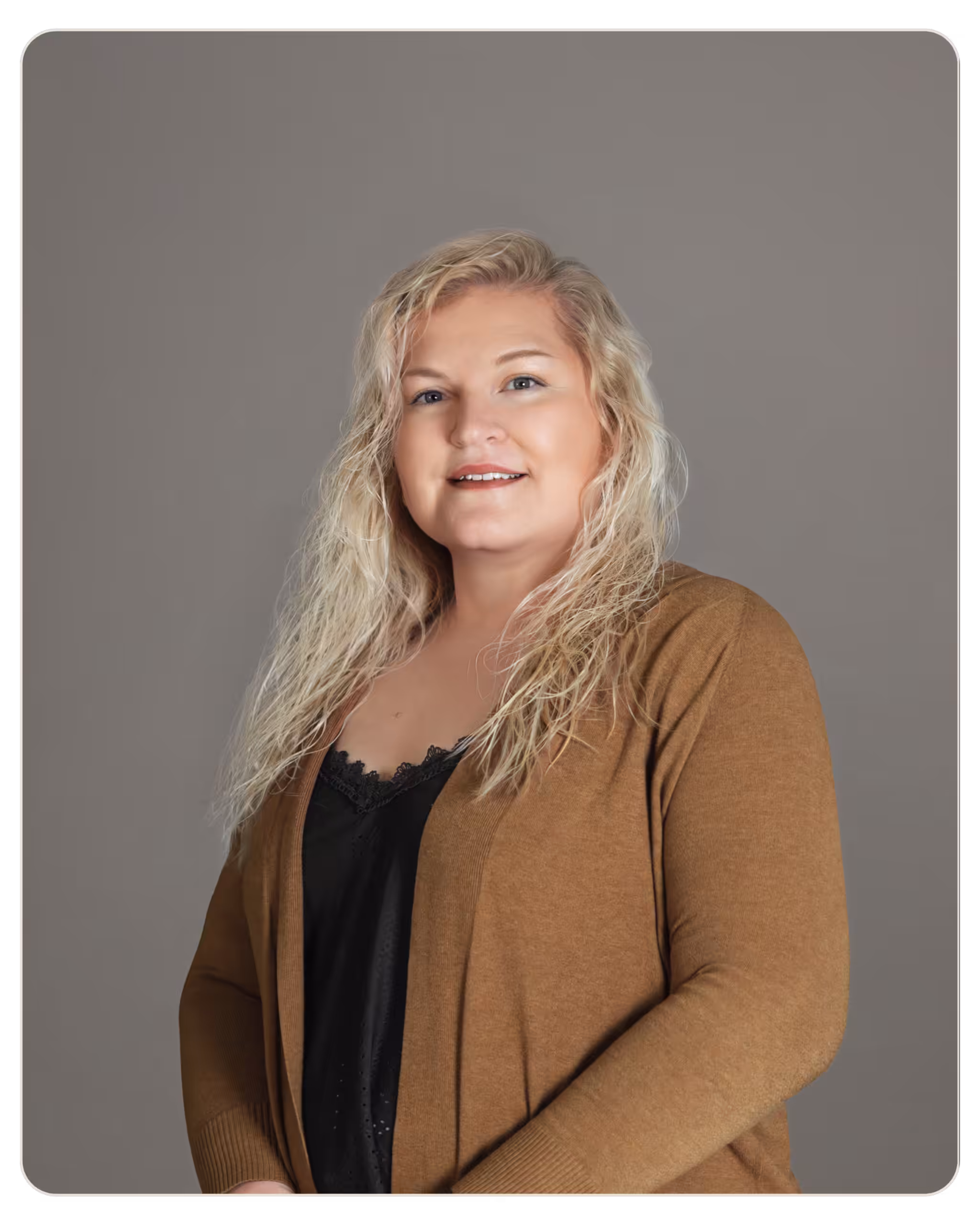 Ember - Cynthia LaPerriere Portrait of a smiling blonde woman in a brown cardigan against a gray studio background with a black lace top visible at the neckline.