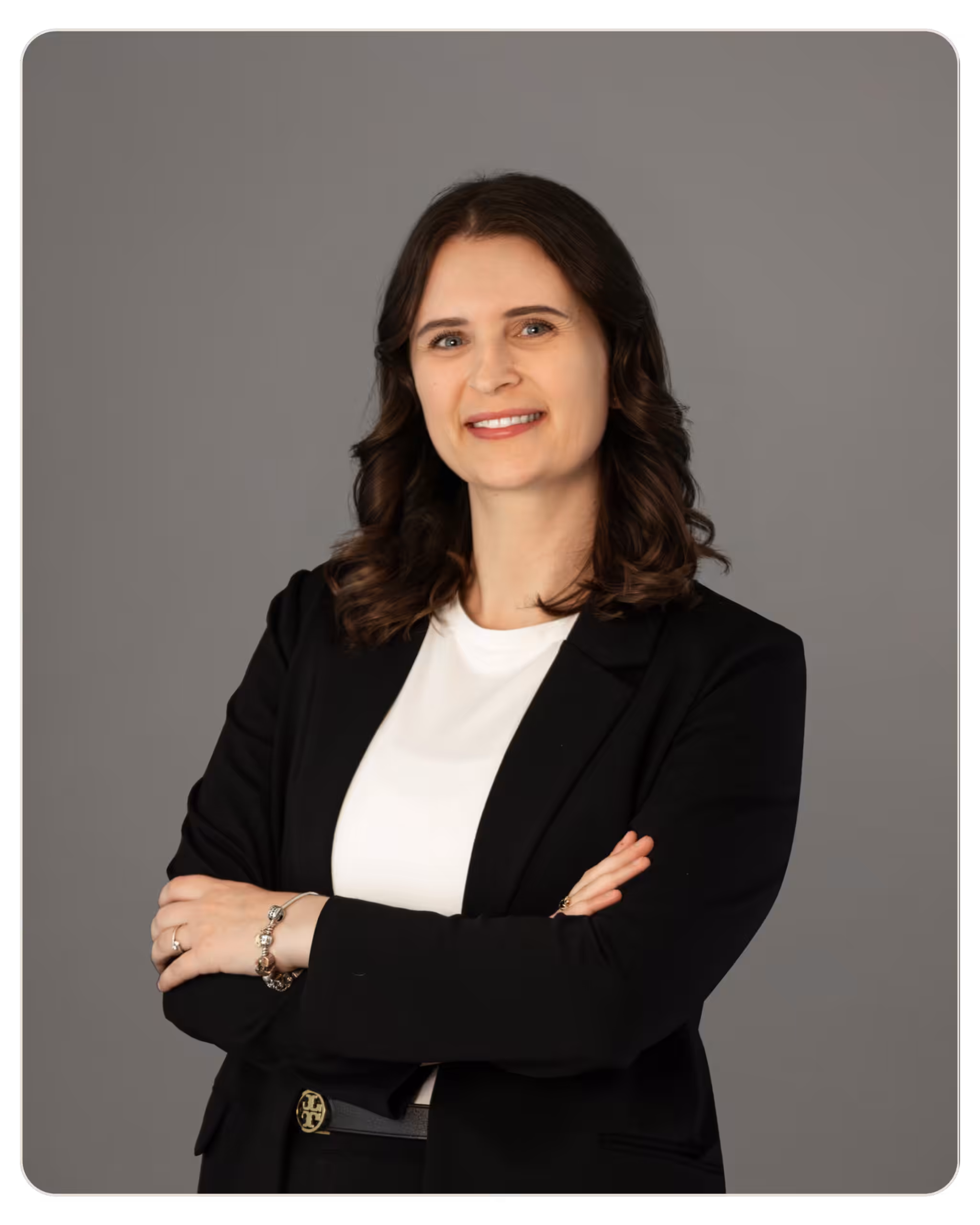 Deanna Stone Kandela Portrait of a smiling businesswoman with arms crossed in a black blazer and white blouse against a gray studio background.