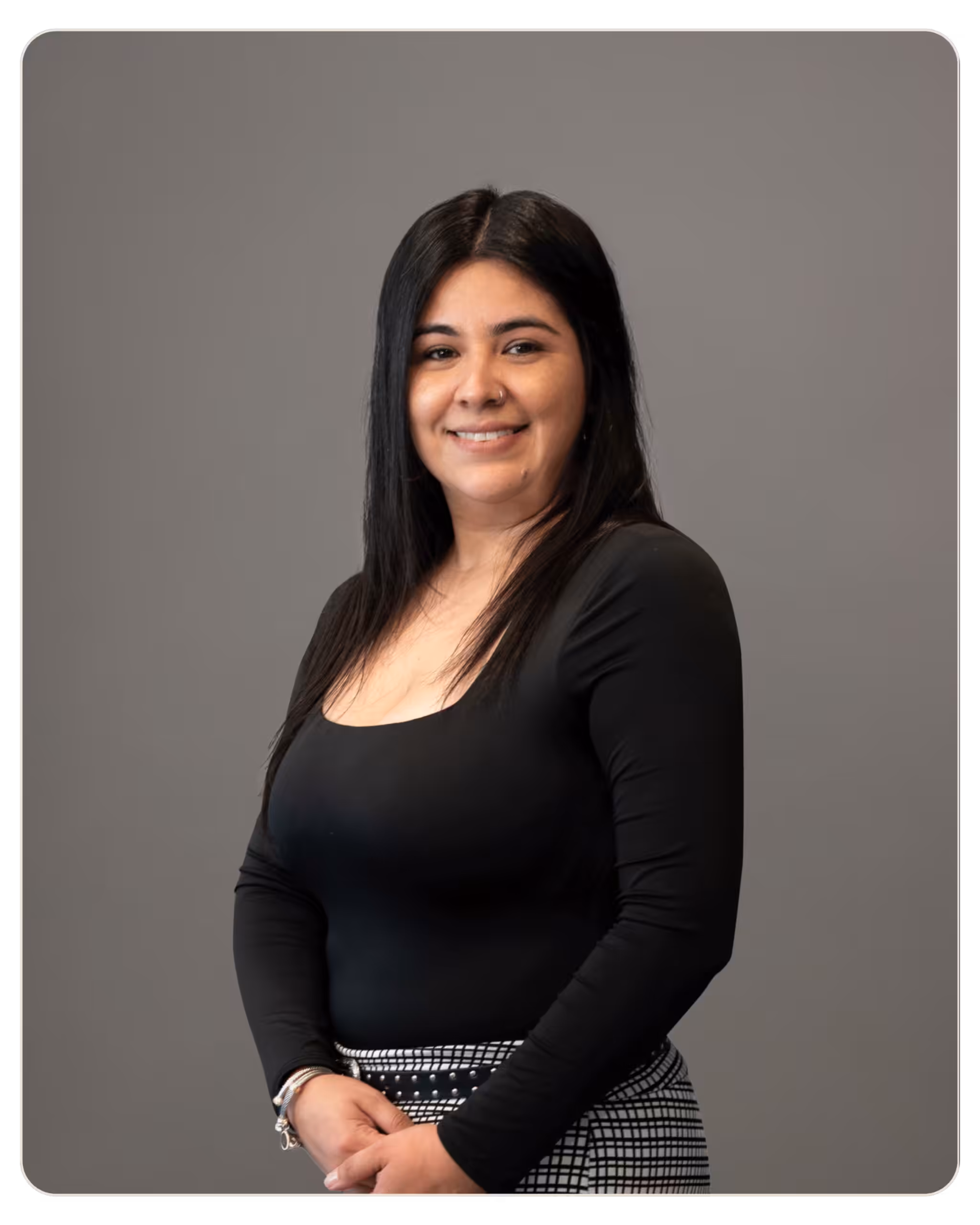 Elizabeth Gonzalez, BS: Case Manager at Aurora. Portrait of a woman with long dark hair, smiling, wearing a black top and black-and-white patterned skirt, against a gray backdrop.