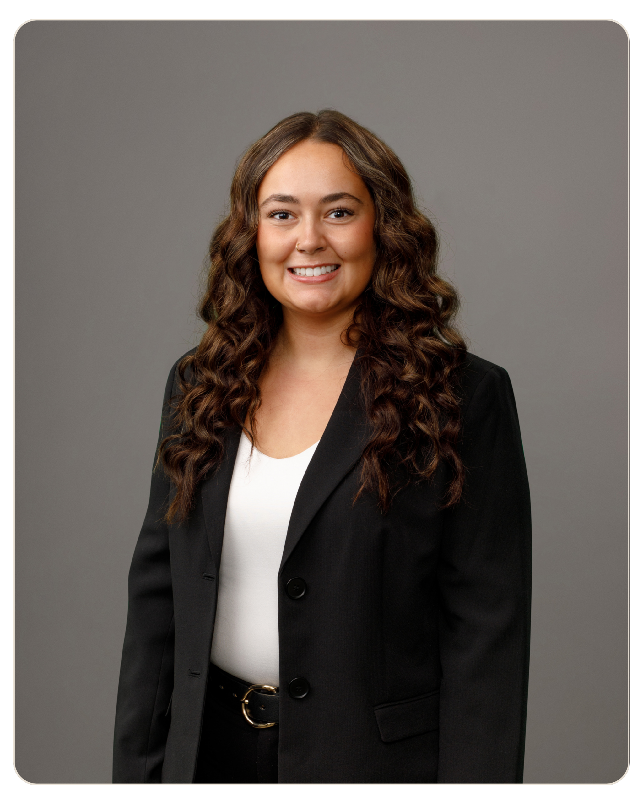 Emma Thrasher Smiling woman with long curly brown hair, wearing a black blazer and white blouse, posed for a professional headshot against a gray backdrop. Smiling woman in a black blazer and white top poses for a studio portrait against a gray backdrop.