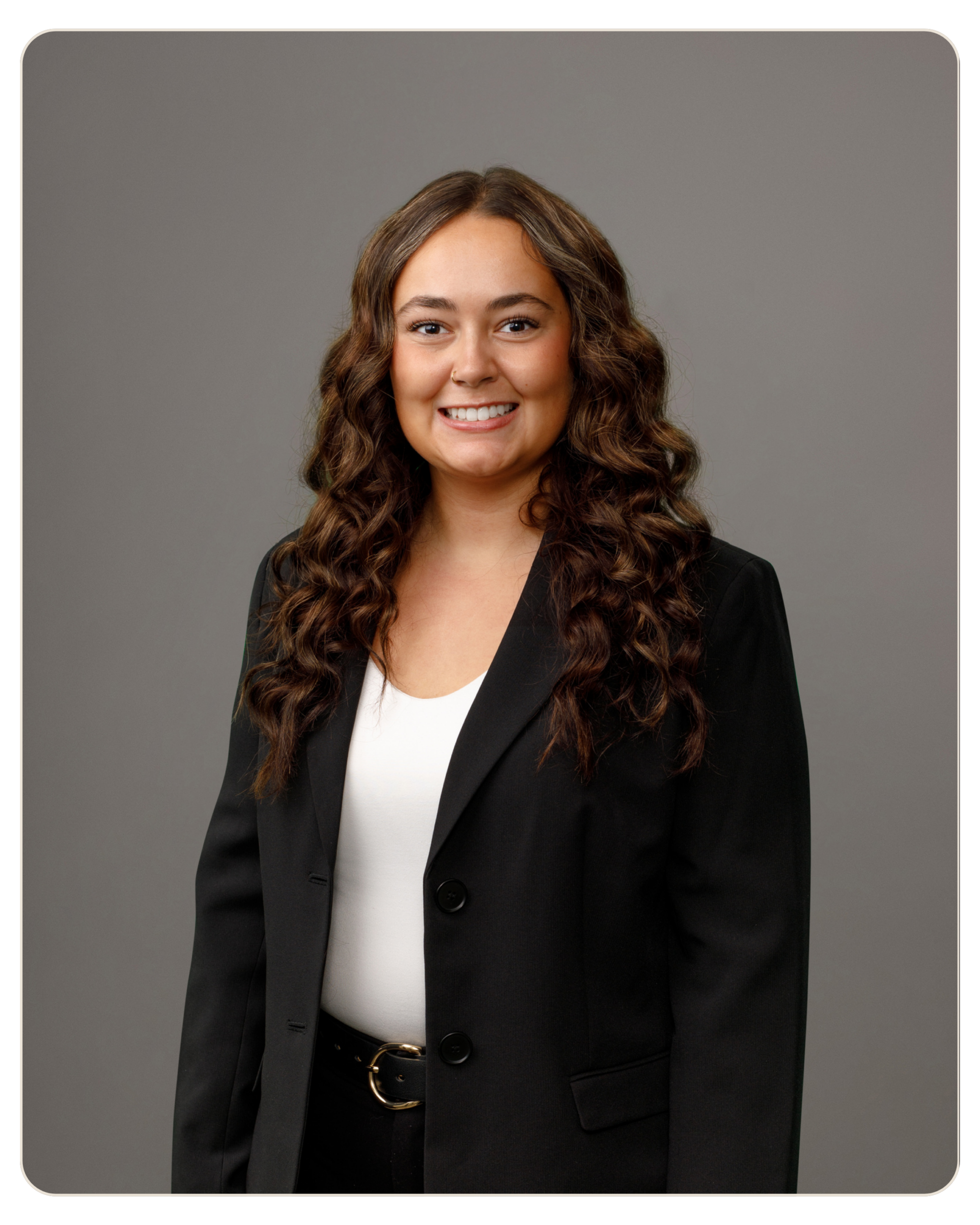 Emma Thrasher Smiling woman with long curly brown hair, wearing a black blazer and white blouse, posed for a professional headshot against a gray backdrop. Smiling woman in a black blazer and white top poses for a studio portrait against a gray backdrop.
