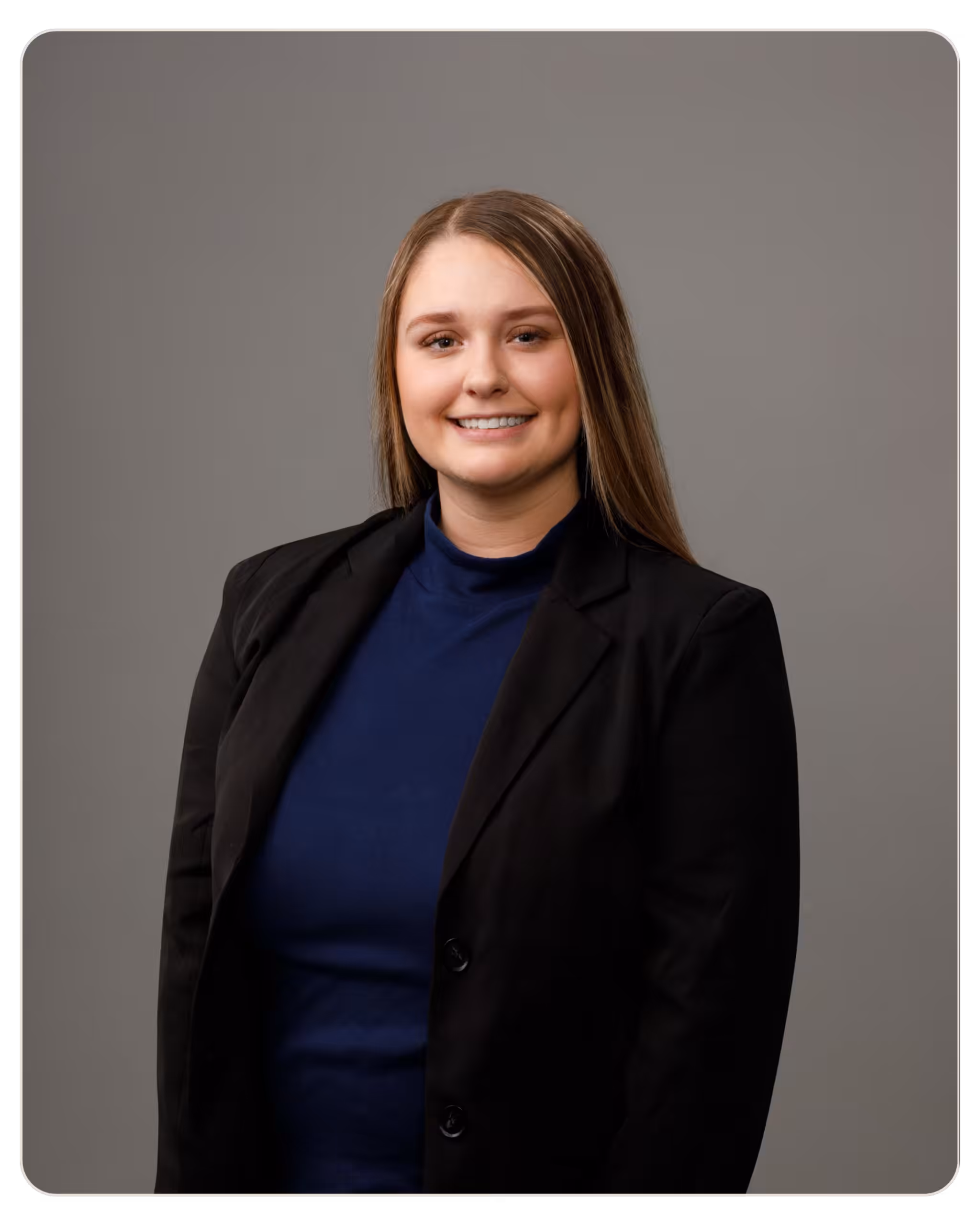 Haile Holcomb Portrait of a smiling woman in a navy top and black blazer against a gray studio backdrop, facing the camera.