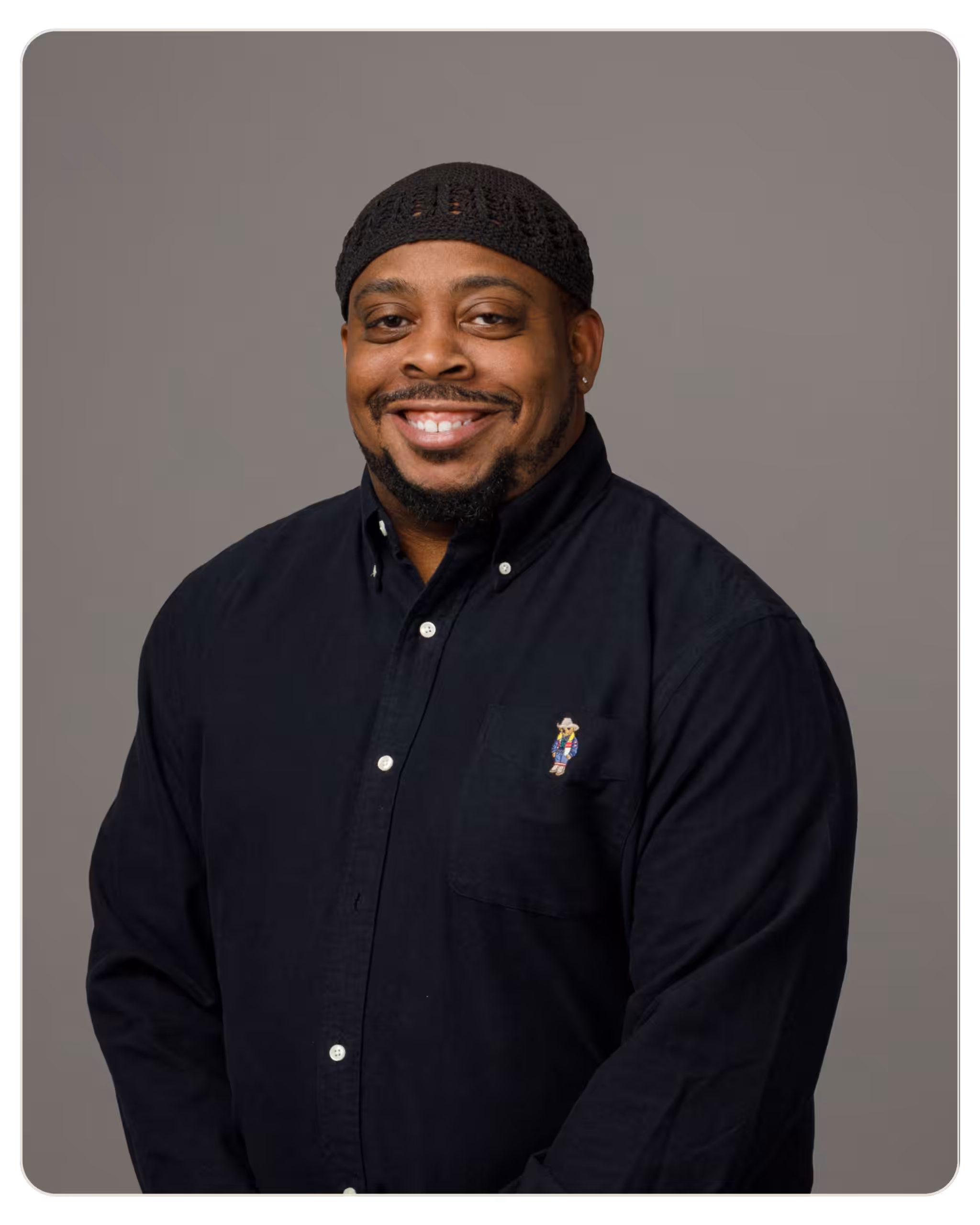 Isiah McCormick: Patient Care Specialist at Beacon. Smiling man wearing a black knit cap and a dark button-down shirt against a gray studio backdrop, portrait-style.