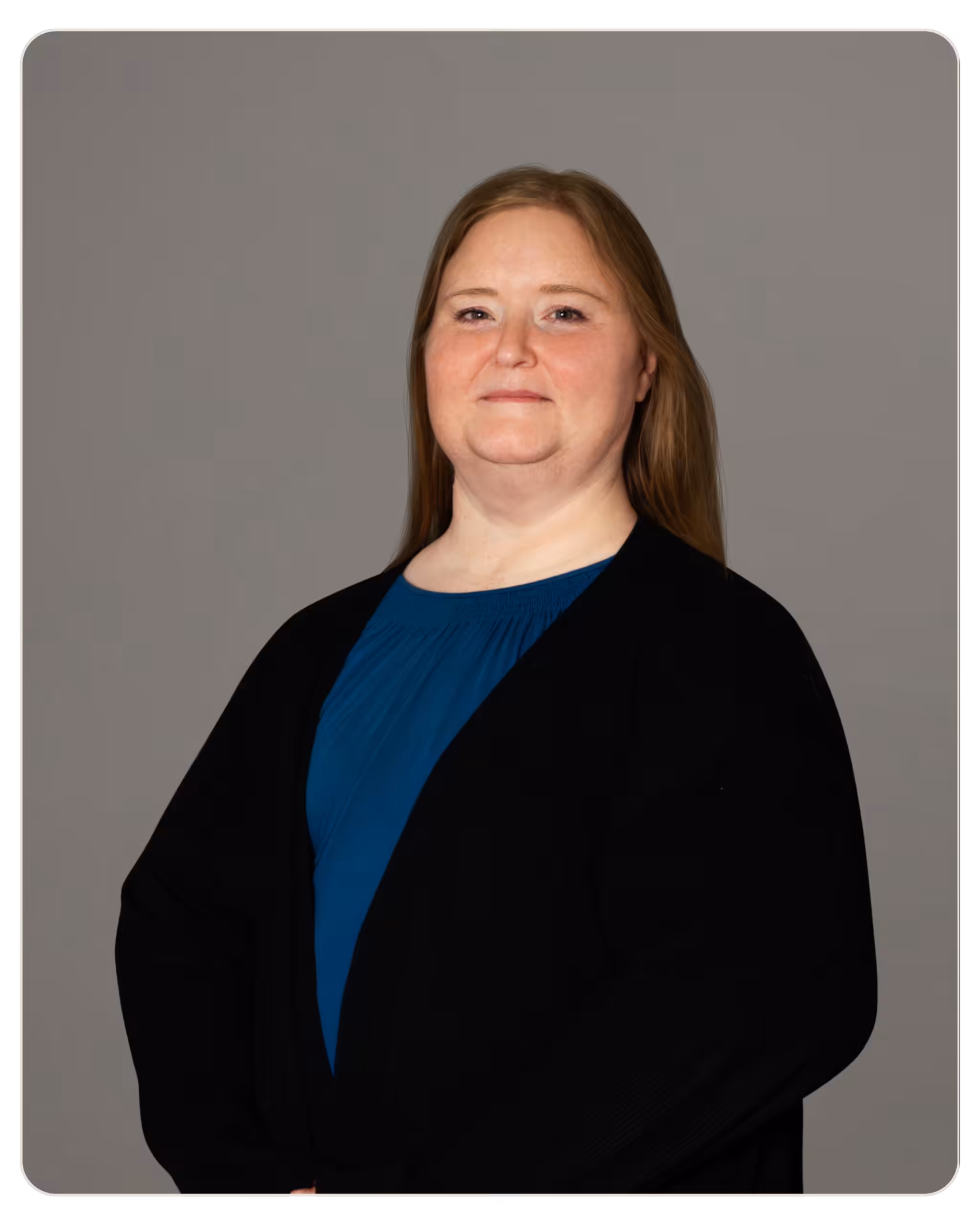 Jodie Simpson, LMFT Portrait of a woman with brown hair wearing a blue blouse and black blazer, arms crossed, against a gray studio backdrop.