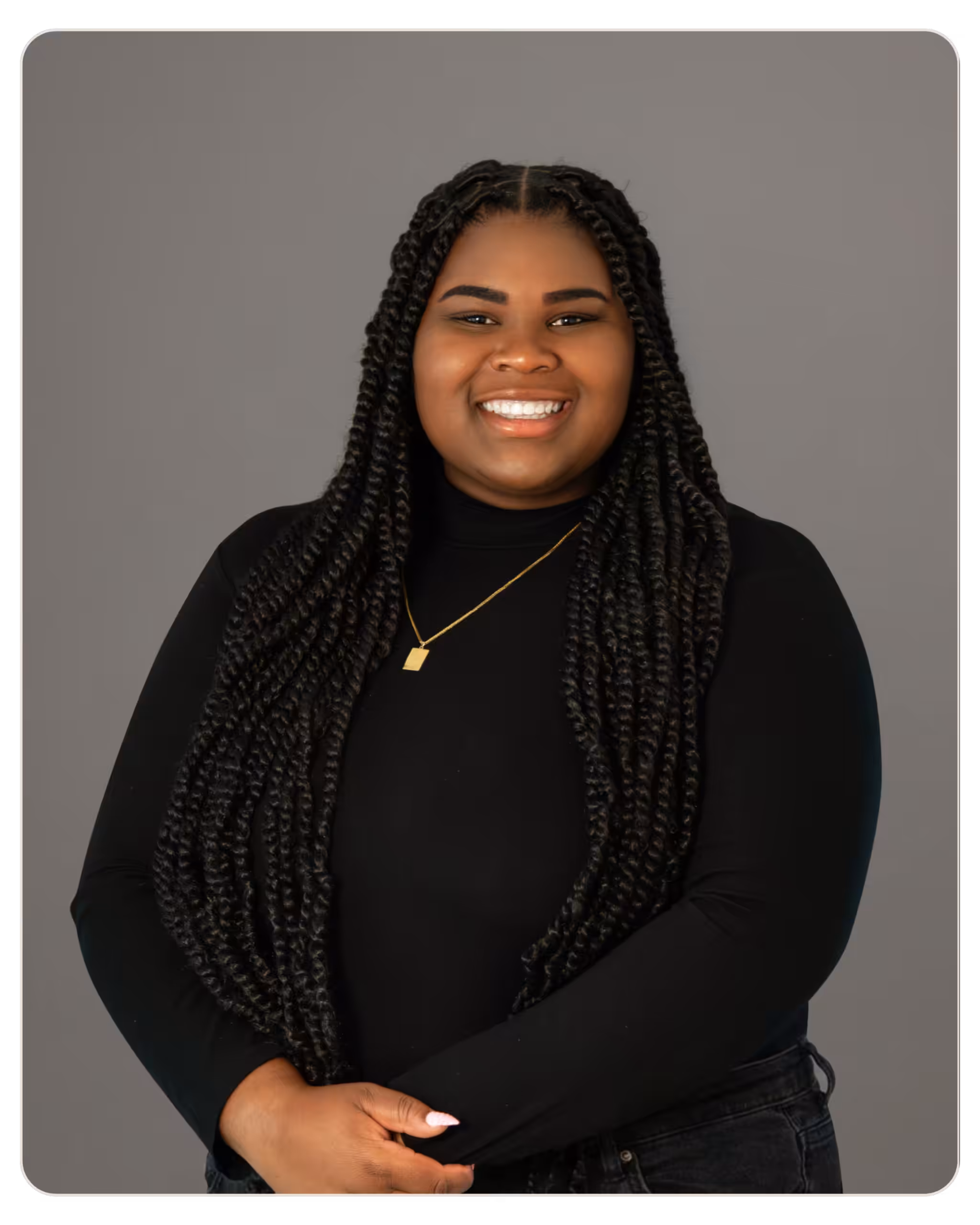 Kayla Waugh RCMHCI Smiling woman with long box braids, wearing a black top and gold necklace against a gray backdrop.