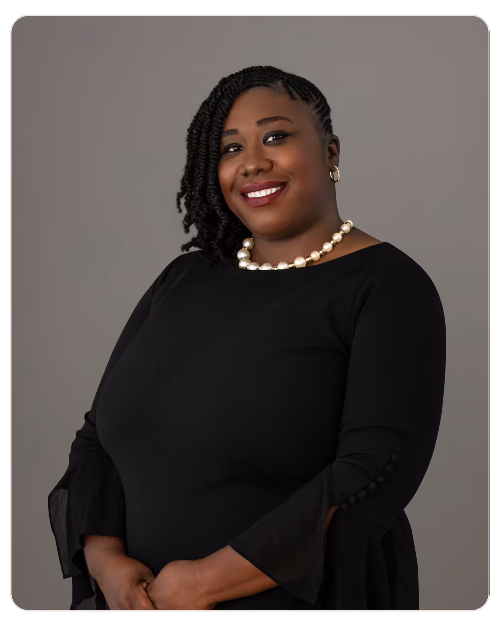 Keshia Frederique, MSW Smiling Black woman in a black dress with a pearl necklace and gold hoop earrings, against a gray background.