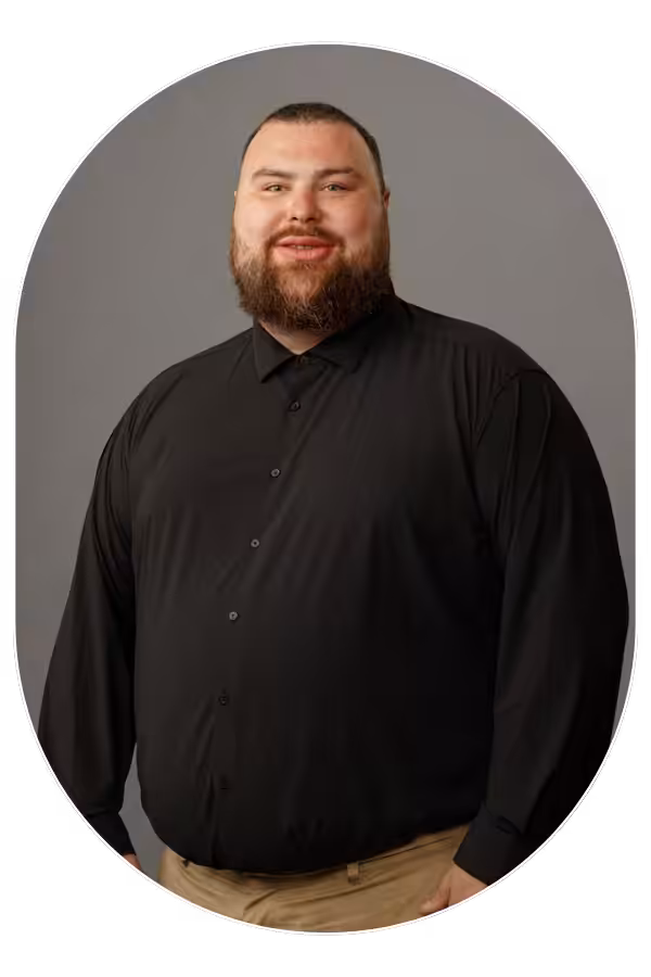 Portrait of a smiling man with a full beard wearing a black button-down shirt, standing against a gray backdrop.