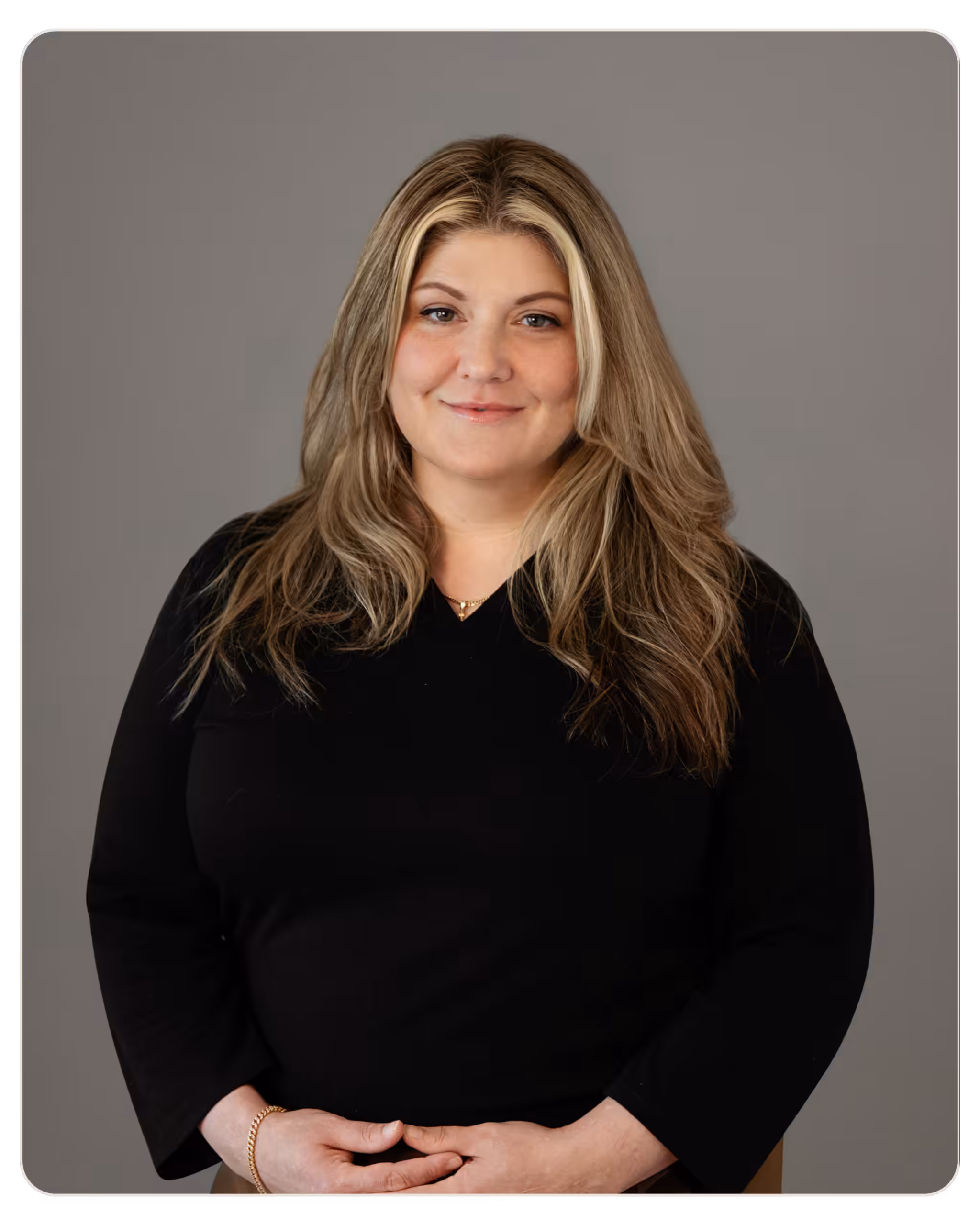Smiling woman with long blonde hair in a black top, hands gently folded in front, gray studio background (profile portrait). Portrait of a smiling woman with long blonde hair and a black top, hands gently clasped in front against a gray background.