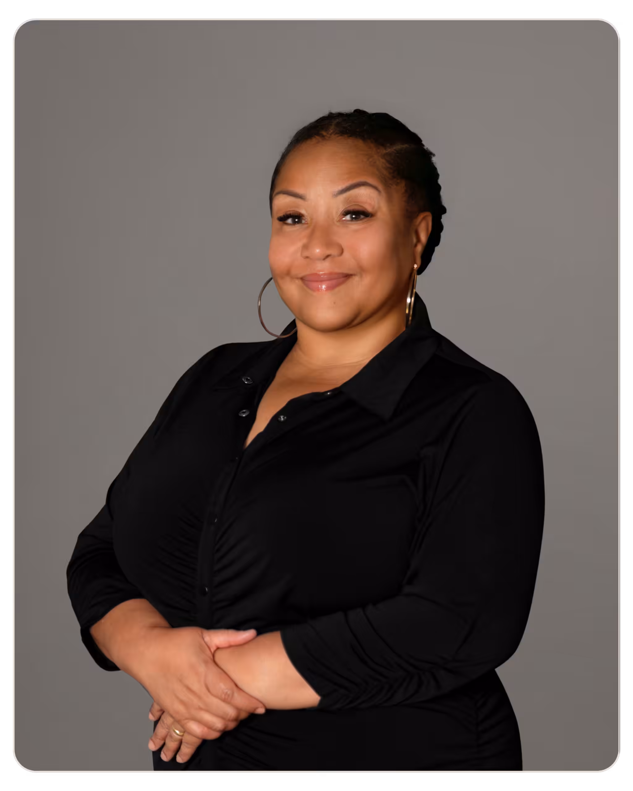 Beacon - Magaly Padilla, MSW Portrait of a smiling woman with braided hair, wearing a black blouse, arms crossed in front of a gray background.