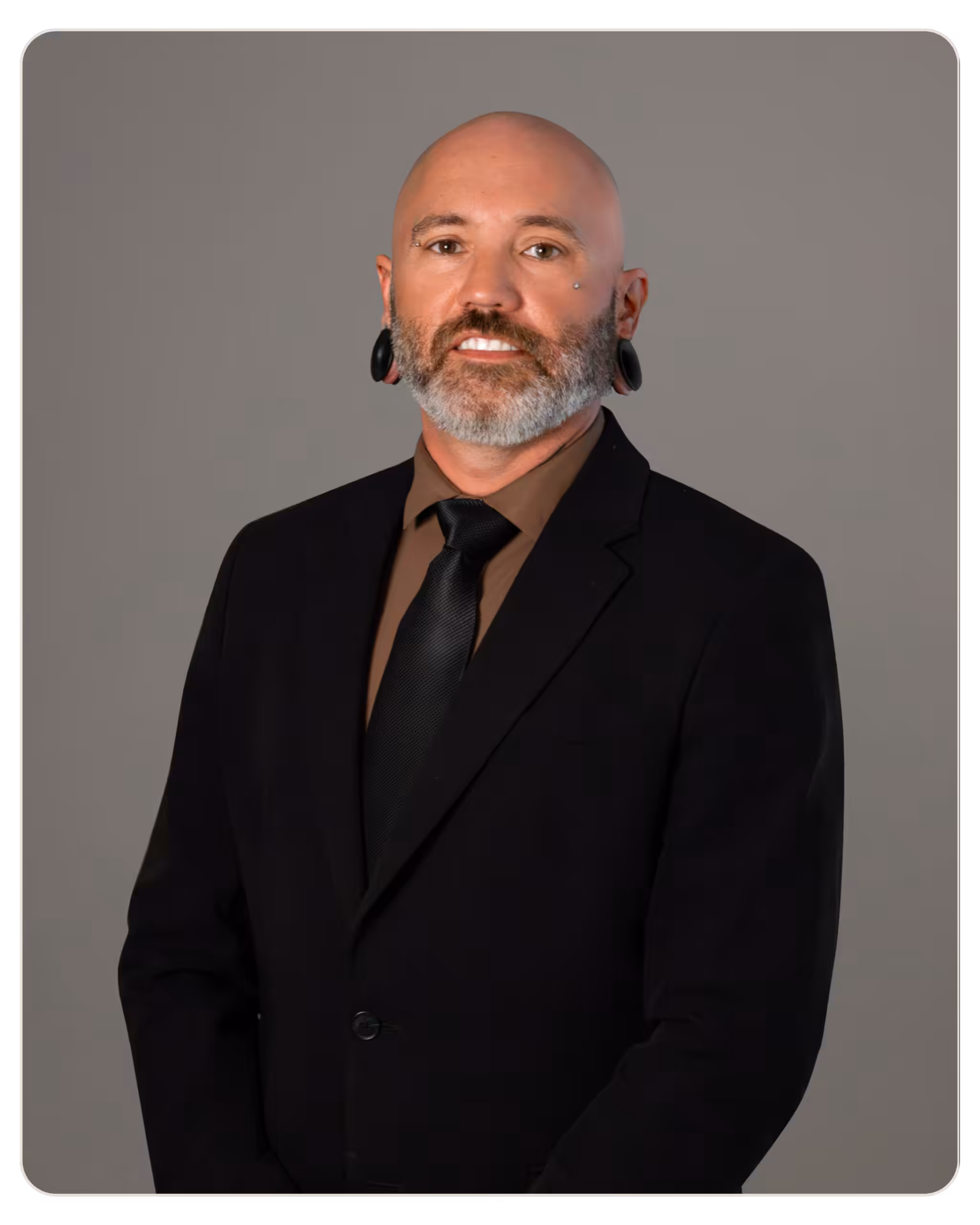 Portrait of a bald man with a gray beard and ear gauges wearing a black suit, brown shirt, and black tie against a gray background.
