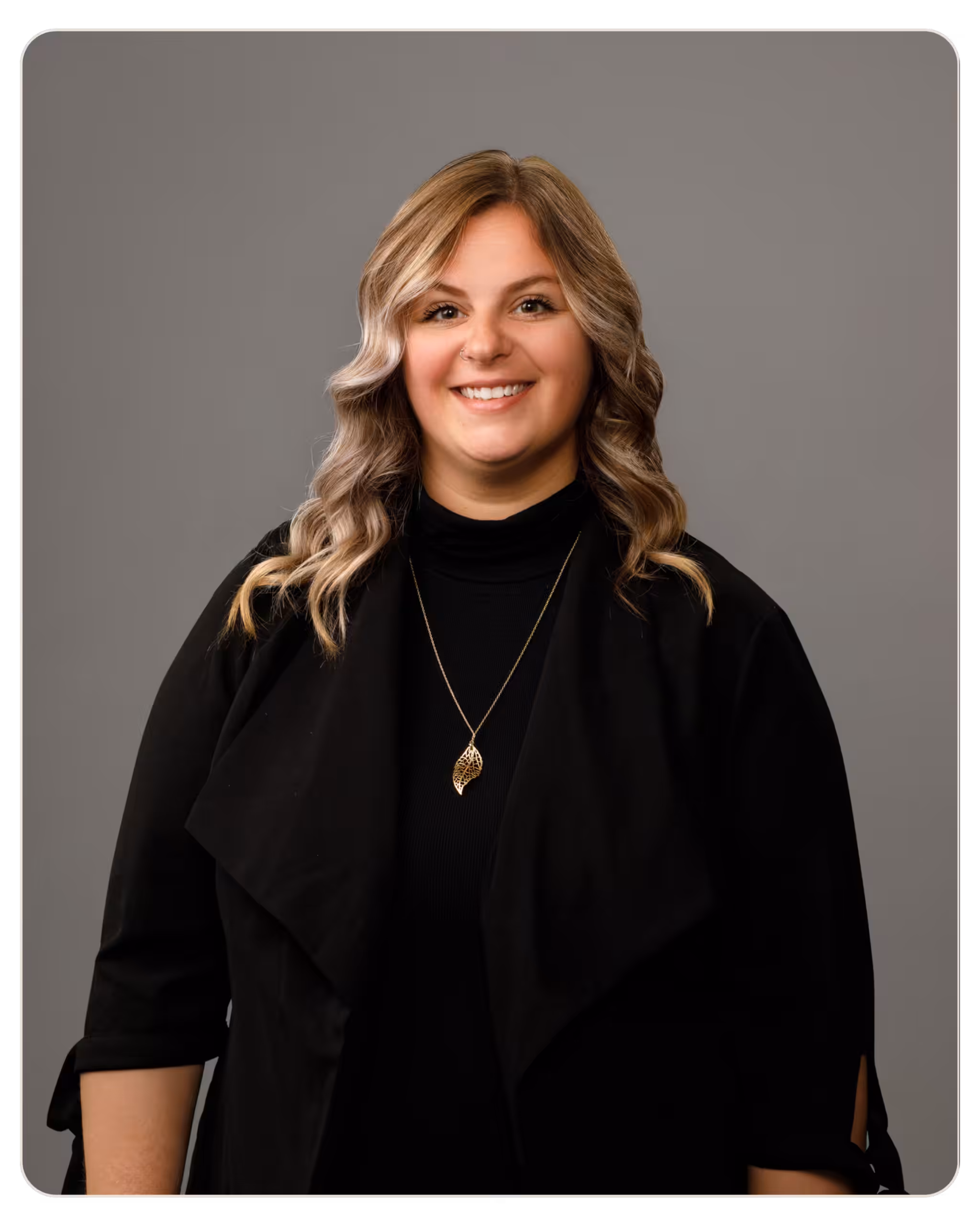 Alexandra Marcus Smiling woman in a black top with a gold necklace, posed against a gray studio background.