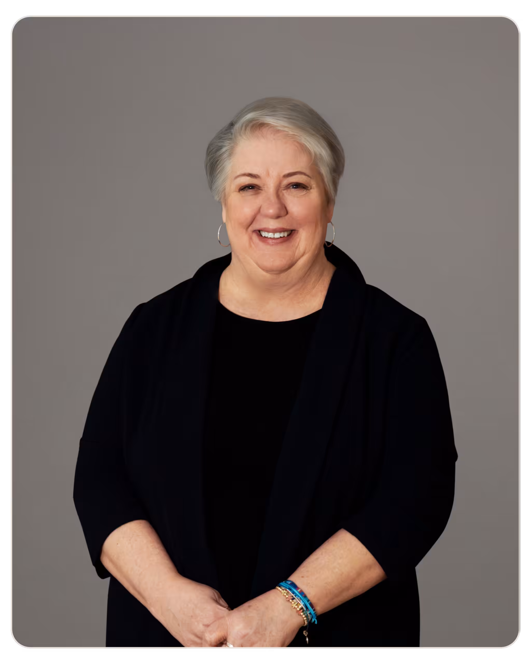 Amy Kitchen - HR Generalist at Skylight Portrait of an older woman with short gray hair, smiling, wearing a black blazer and hoop earrings against a gray backdrop.