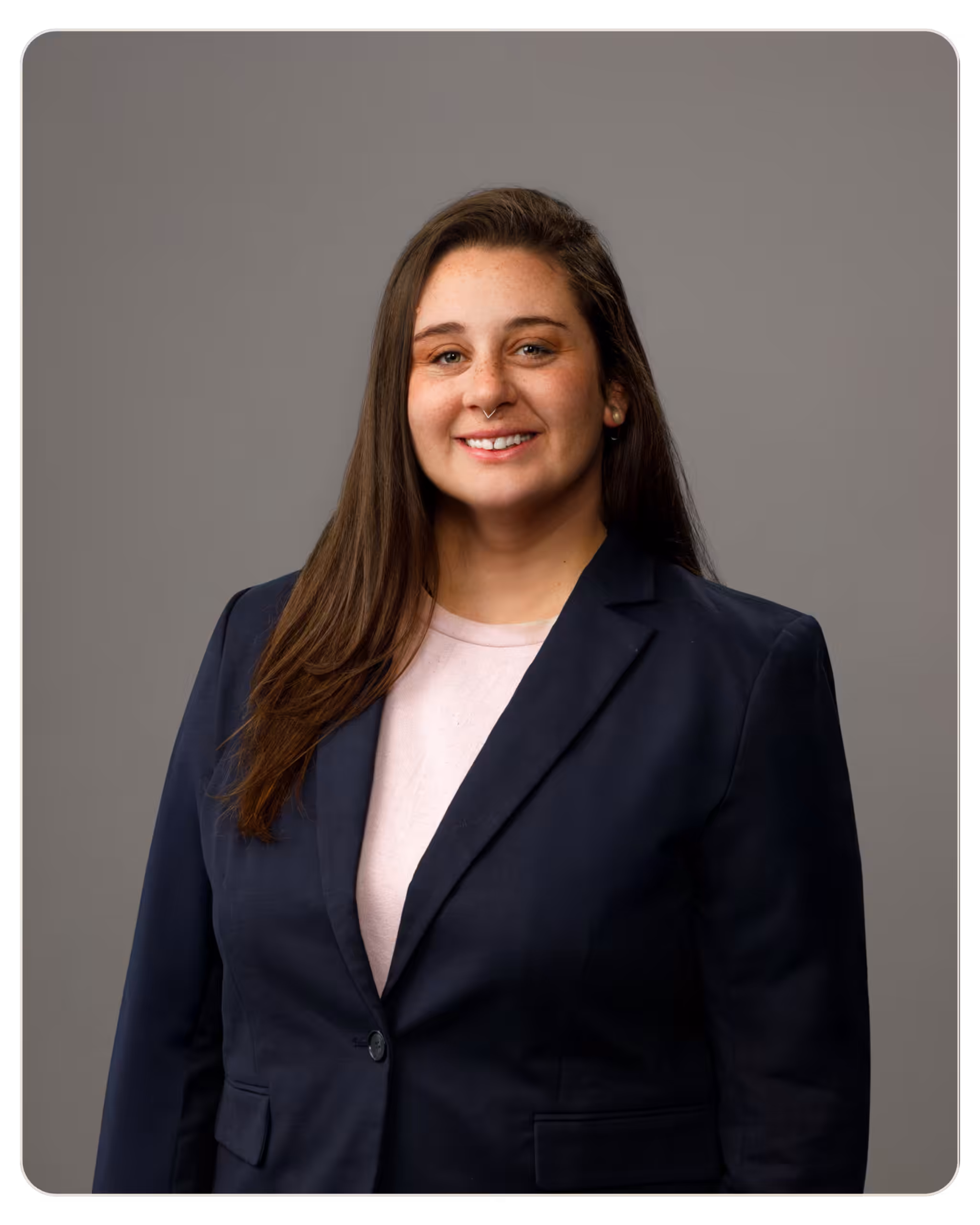 Skylight - Carlynne Jones Portrait of a smiling woman with long brown hair wearing a navy blazer over a pale pink top against a gray studio background, in a professional head-and-shoulders shot.
