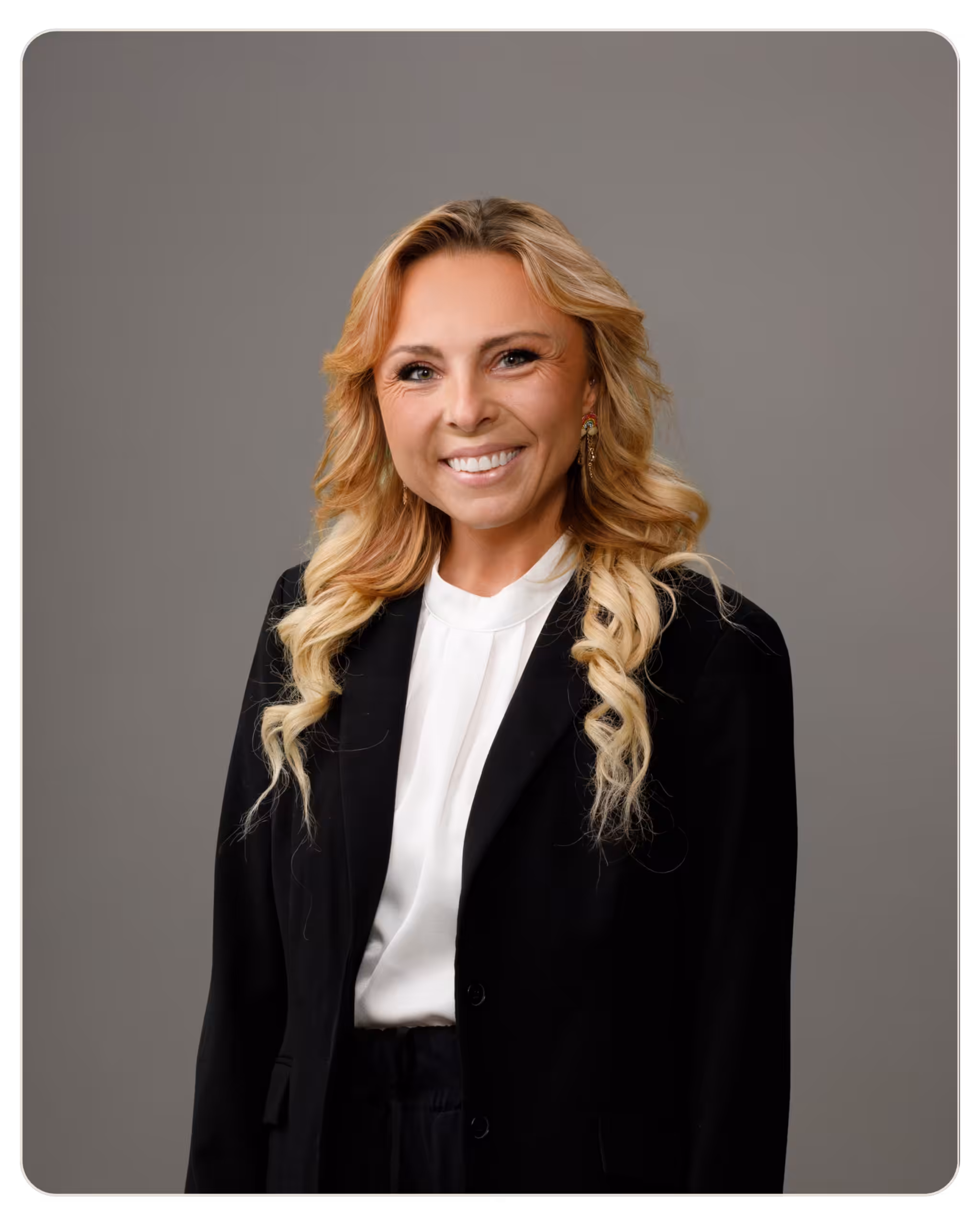 Elizabeth VanSant Smiling blonde woman in a black blazer and white blouse, studio headshot against a gray background.