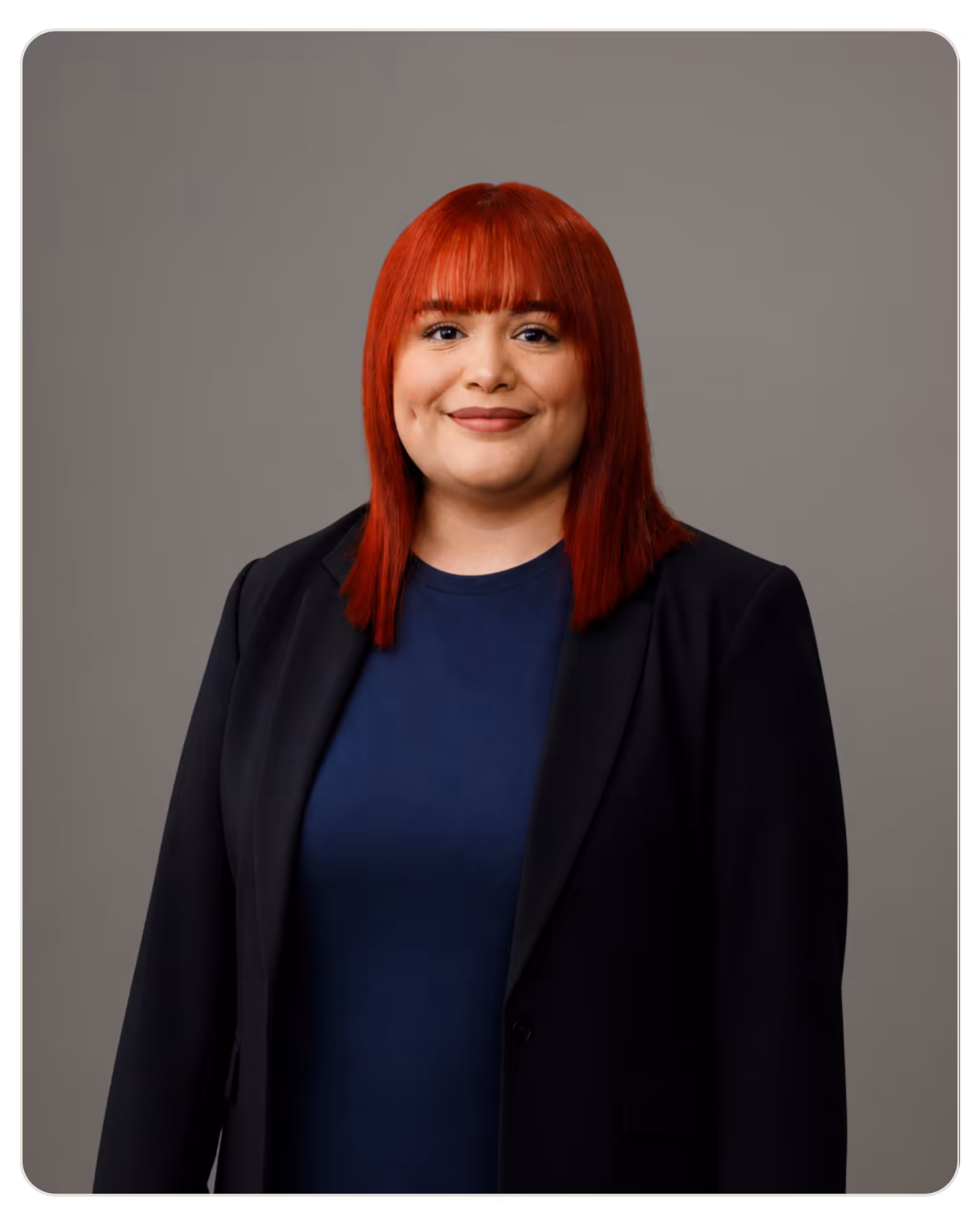 Keila Hernandez Head-and-shoulders portrait of a woman with bright red hair, wearing a navy top and black blazer, against a gray backdrop