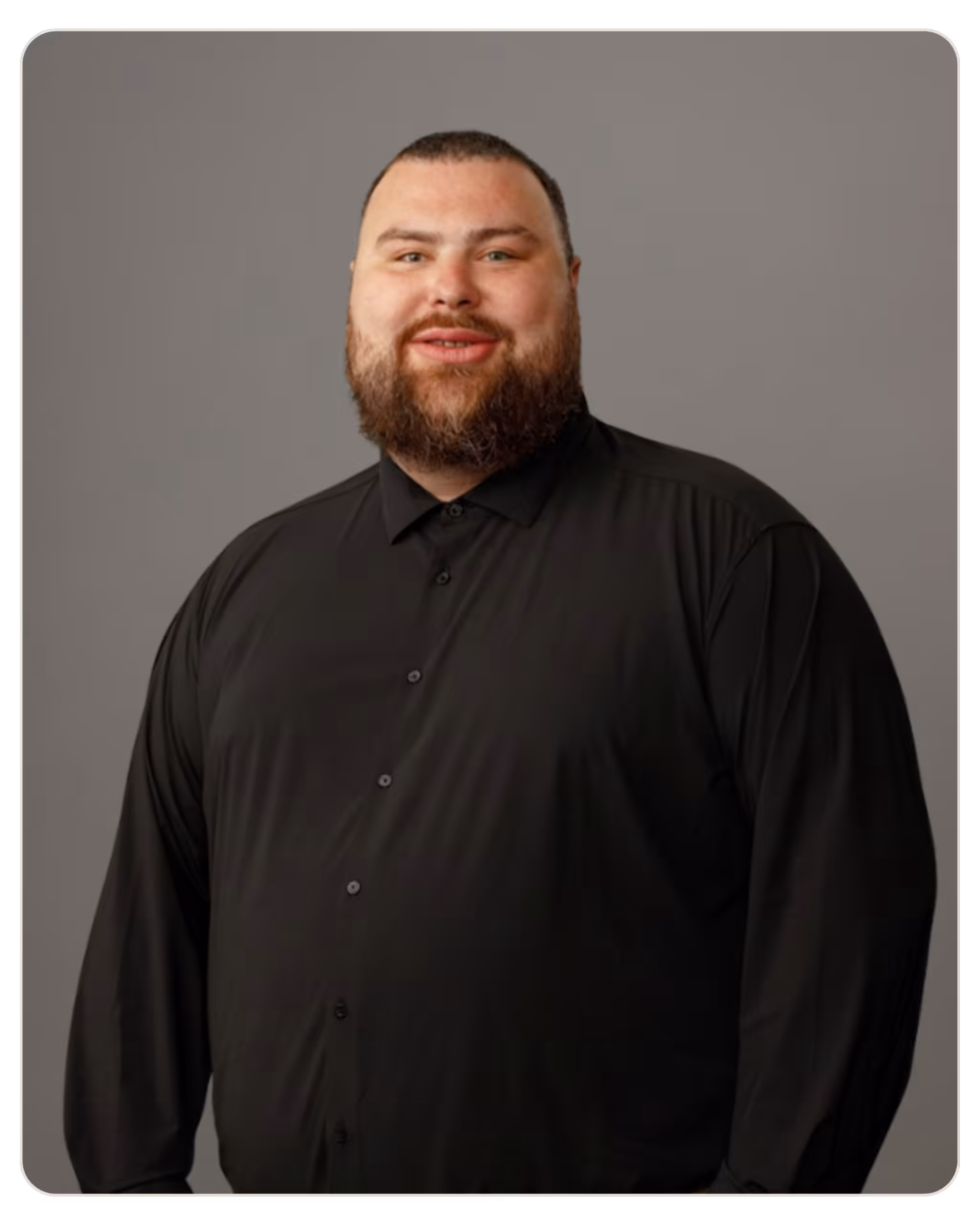 Portrait of a smiling man with a full beard wearing a black button-down shirt, standing against a gray backdrop. Portrait of a man with a short beard and dark hair, wearing a black button-down shirt against a gray backdrop.