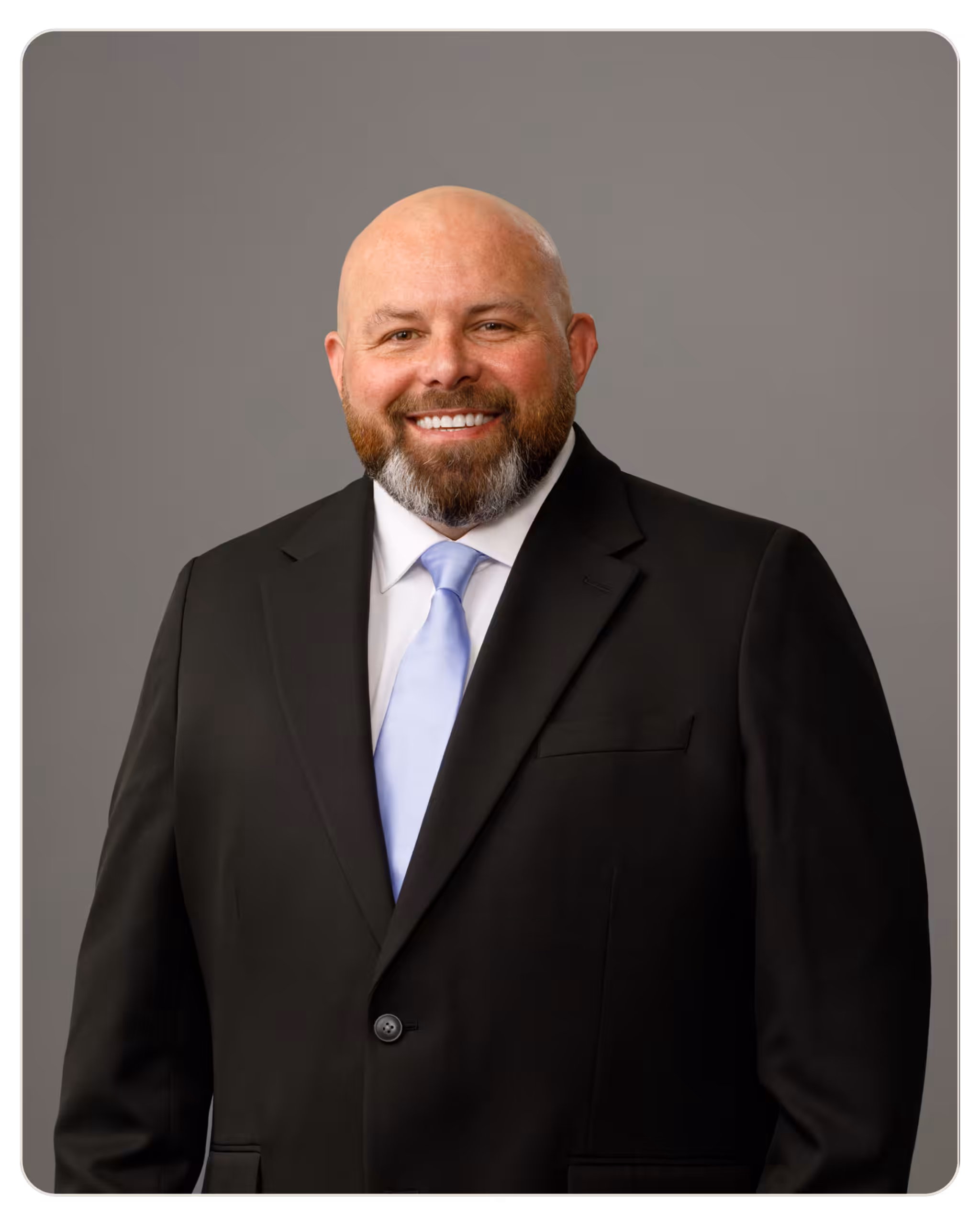 Mike Captain Smiling bald man with a beard in a black suit and light blue tie, posing against a gray backdrop.