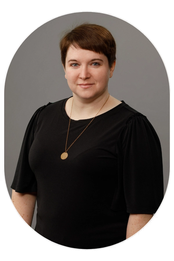 Portrait of a woman with short brown hair wearing a black blouse and gold pendant necklace, gray background.