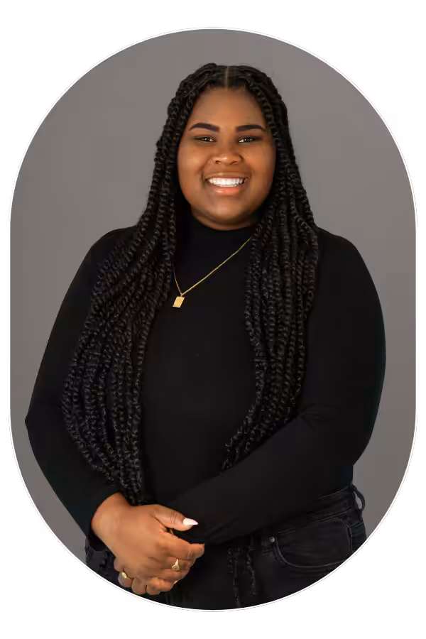 Smiling woman with long box braids wearing a black top and gold necklace, standing against a gray backdrop.