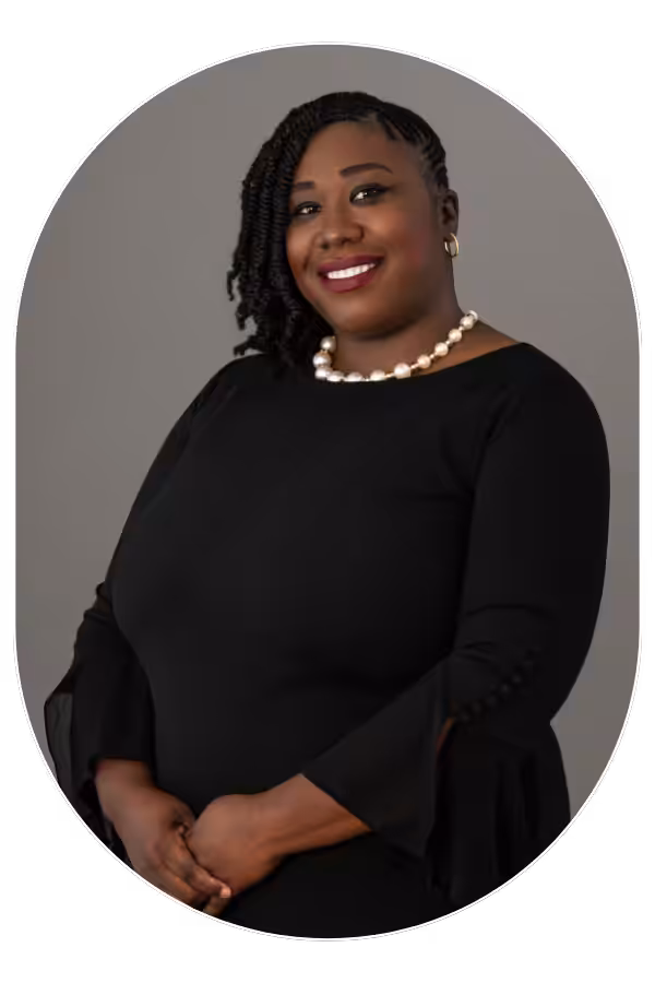 Portrait of a smiling Black woman in a black dress with a pearl necklace, against a gray background.