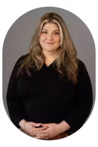 Smiling woman with long blonde hair in a black top, hands gently folded in front, gray studio background (profile portrait).