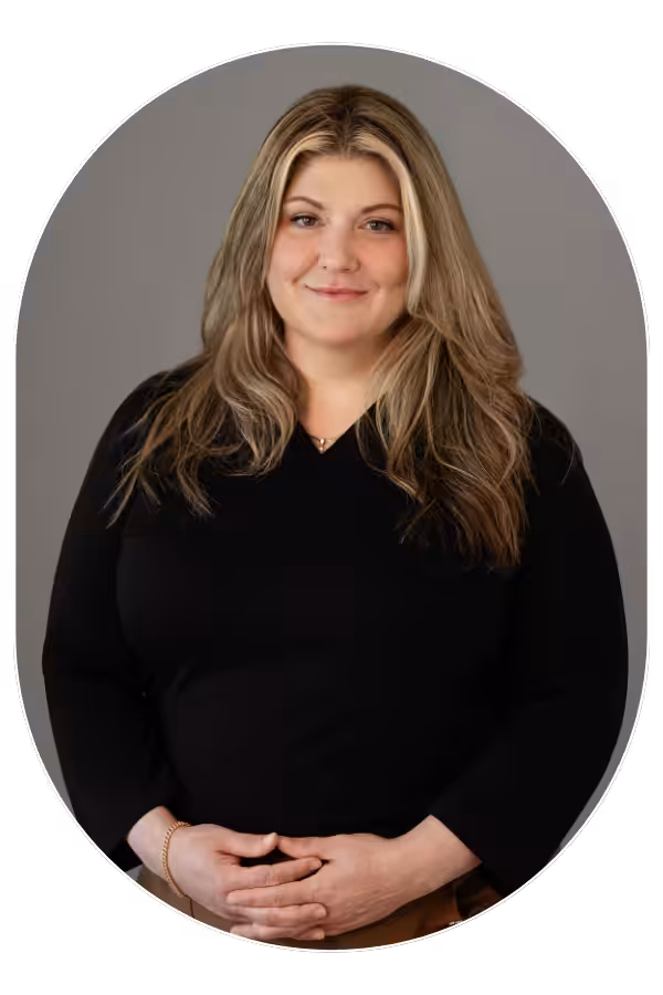 Smiling woman with long blonde hair in a black top, hands gently folded in front, gray studio background (profile portrait).
