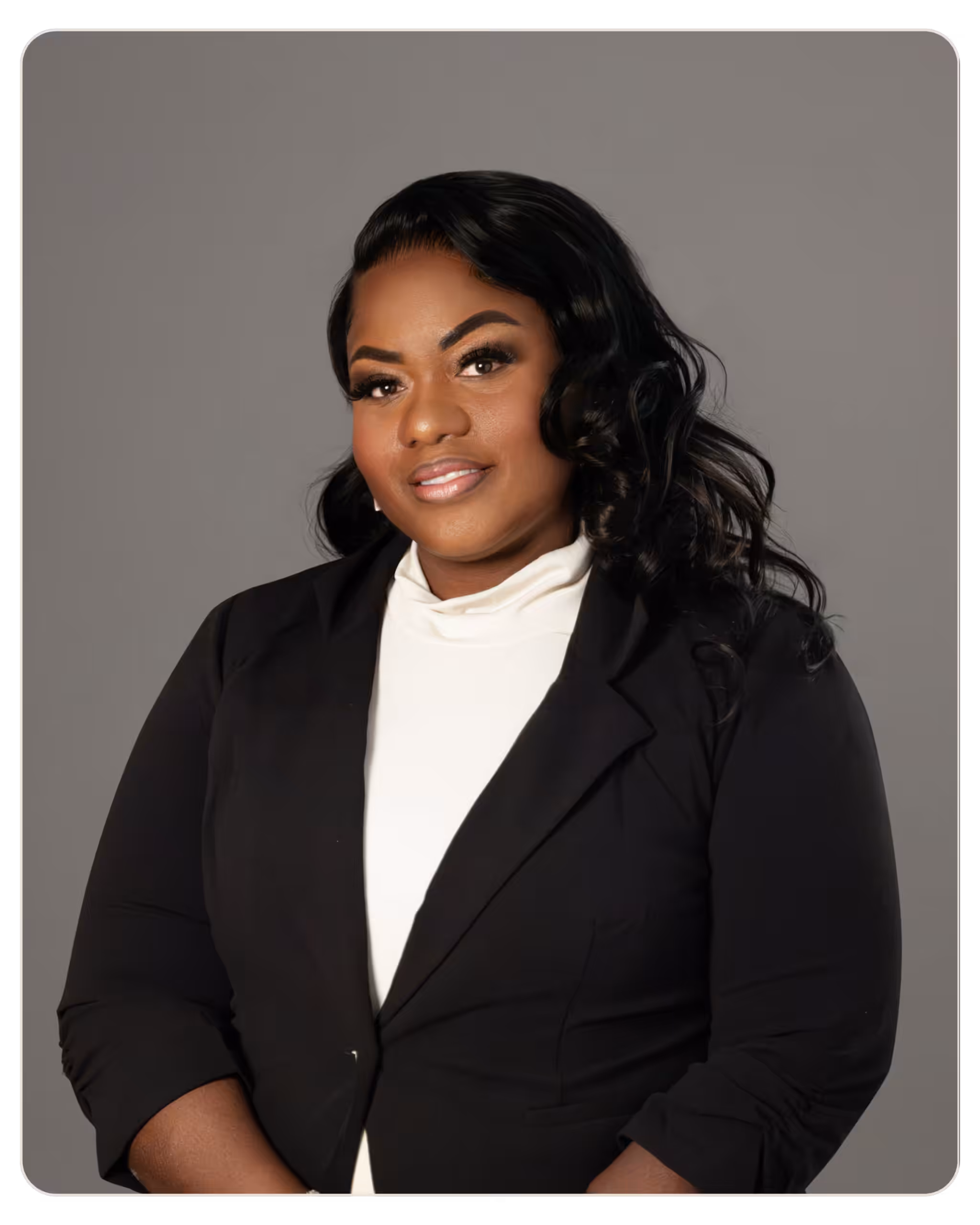 Anishia Wright - Daylight Professional headshot of a smiling woman with black hair, wearing a black blazer and white blouse against a gray background in a studio setting.