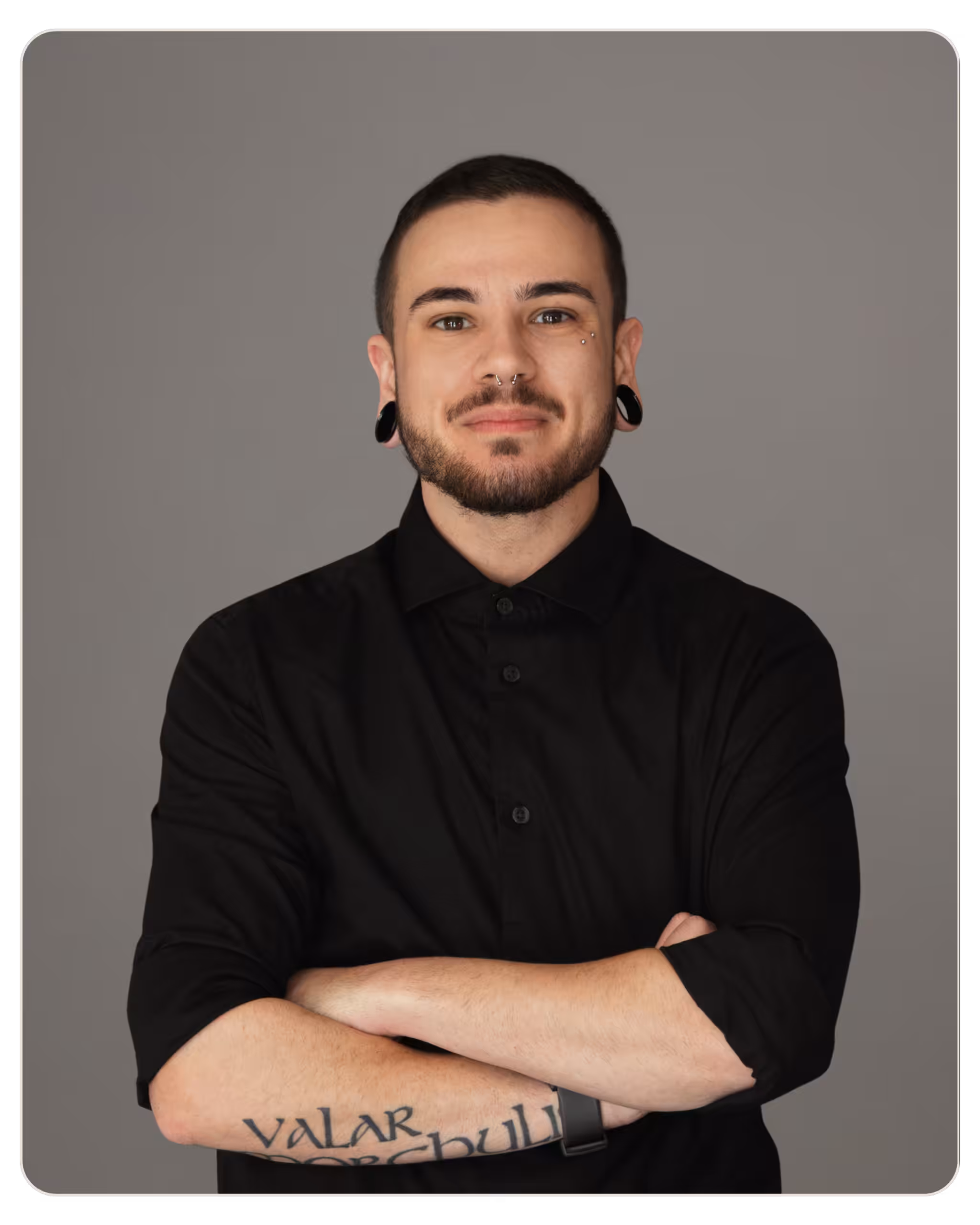 Bennett Willis Portrait of a man with facial piercings and gauge earrings, wearing a black shirt, arms crossed against a gray background.