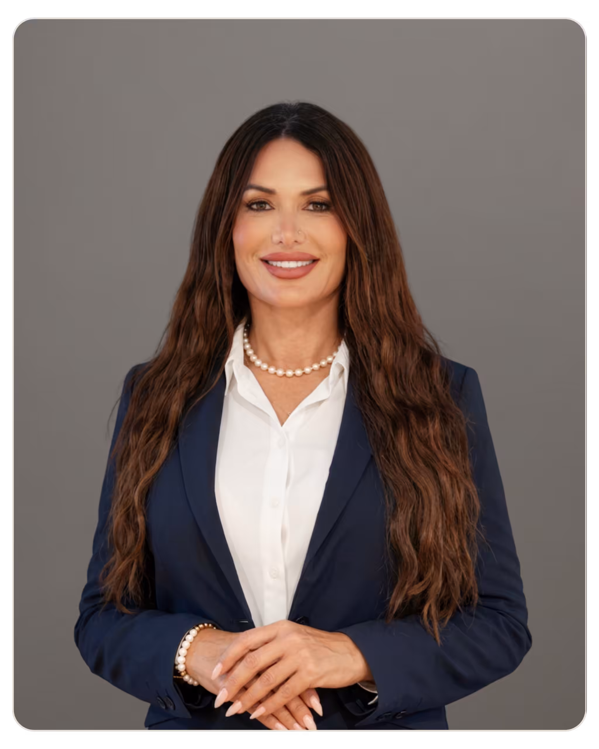 Carol Roldan, LMHC, CFMT, Q-S, Prisma Portrait of a professional woman with long brown hair, wearing a navy blazer, white blouse, and a pearl necklace, smiling with folded hands in front.