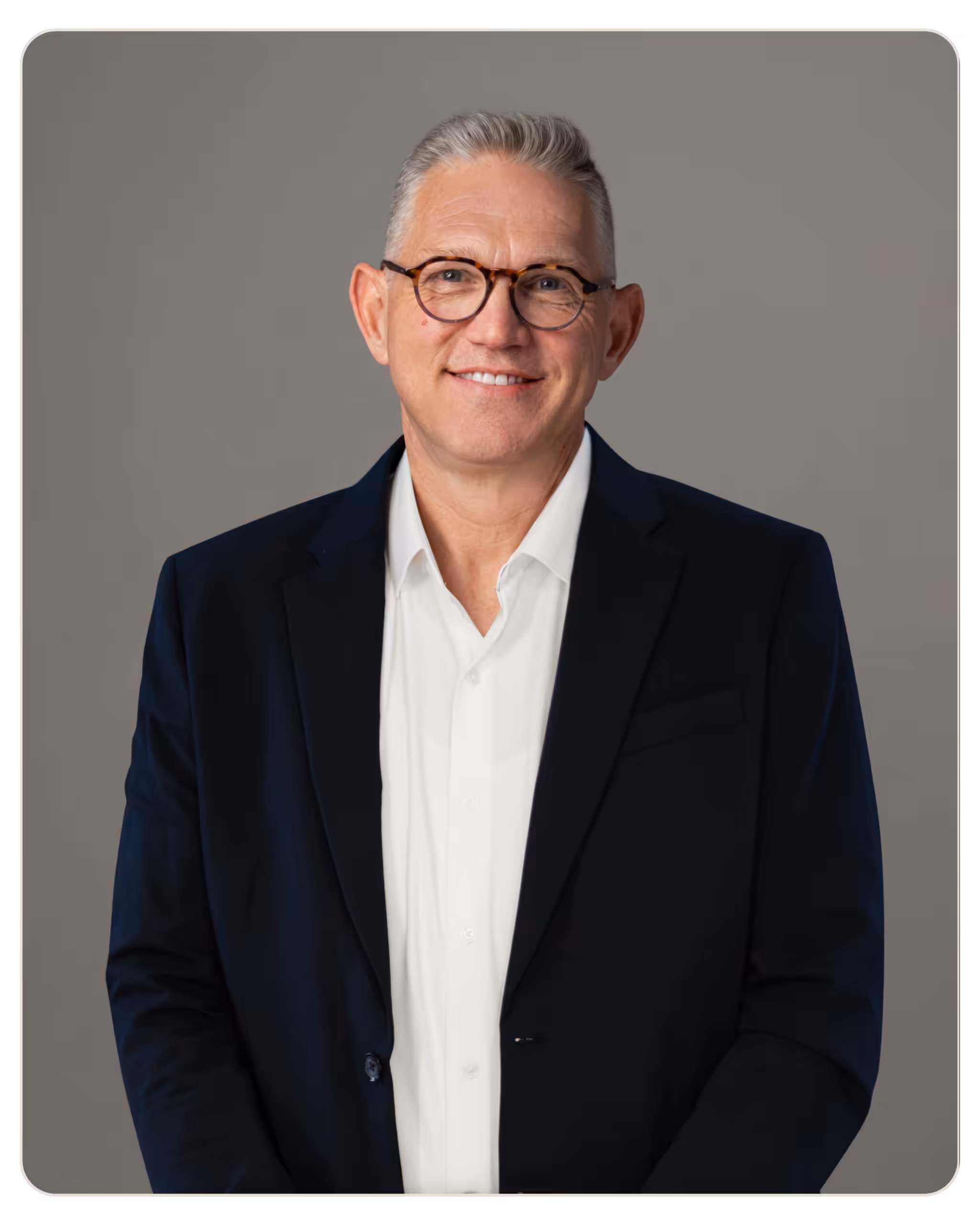 Portrait of a smiling middle-aged man in a dark suit and white shirt, wearing glasses. Smiling man in a navy blazer and white shirt wearing round tortoiseshell glasses, gray studio backdrop.