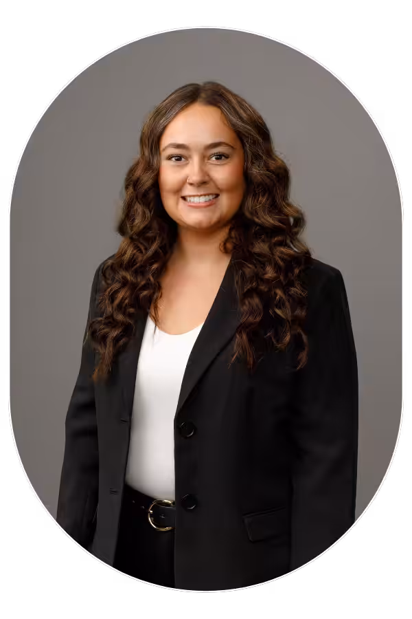 Smiling woman with long curly brown hair, wearing a black blazer and white blouse, posed for a professional headshot against a gray backdrop.