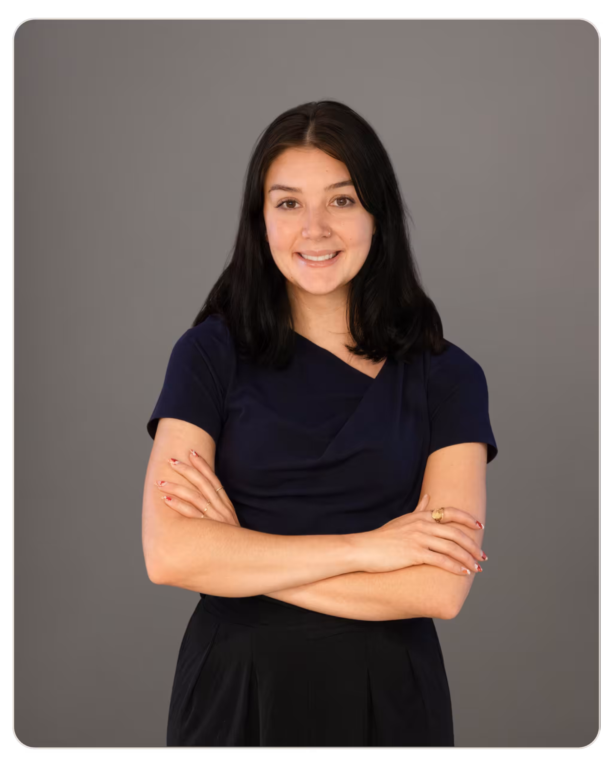Smiling woman with shoulder-length dark hair in a navy top and black skirt, posed against a gray backdrop. Smiling woman with dark hair wearing a navy top, arms crossed, against a gray backdrop.