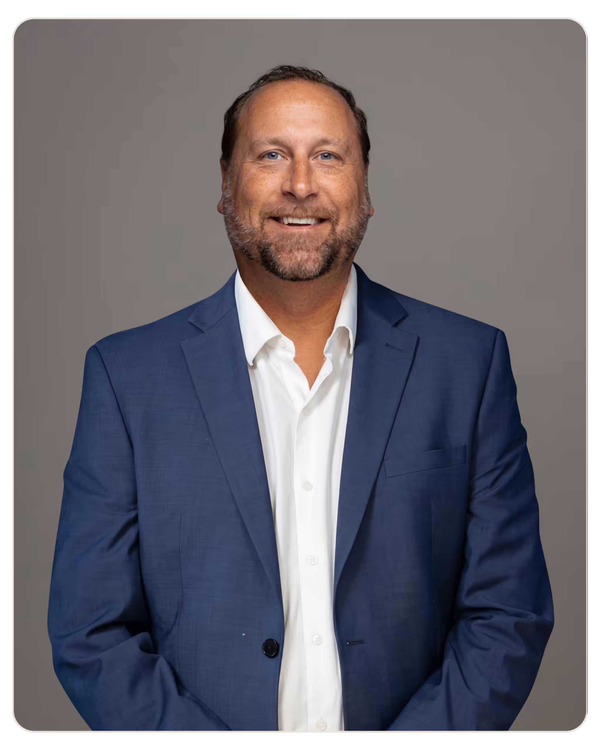 Matthew Lee, Director of Operations at Prisma Recovery Center. Portrait of a smiling man in a navy suit and white shirt against a gray background, in a rounded portrait frame, full torso view.