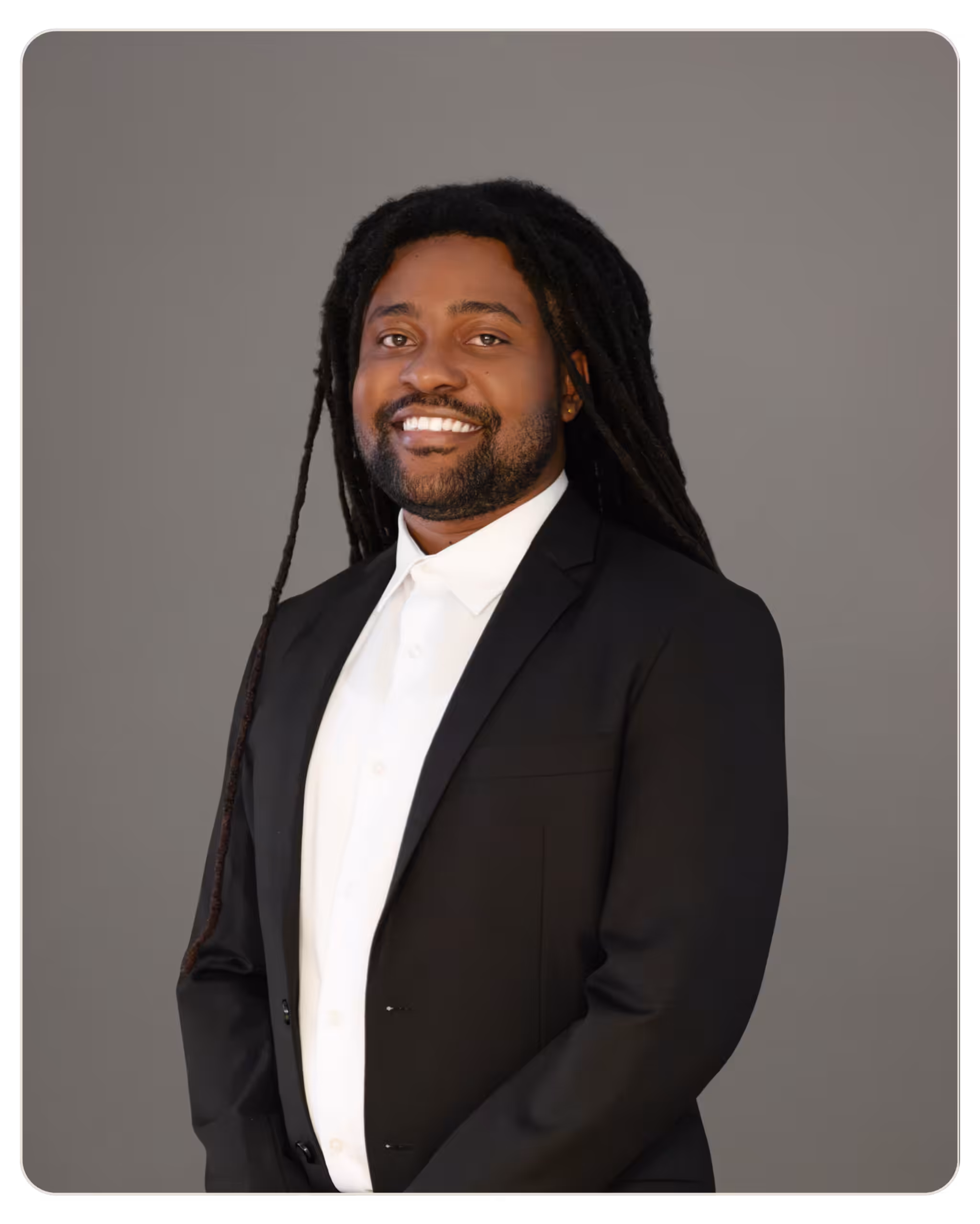 Smiling man with long dreadlocks in a black suit and white shirt, standing against a gray backdrop. Smiling man with dreadlocks in a black suit and white shirt, posed for a formal portrait.