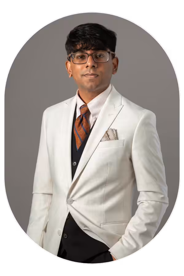 Portrait of a man wearing a white blazer, dark vest, orange patterned tie, and glasses, posing against a gray background with a pocket square showing.