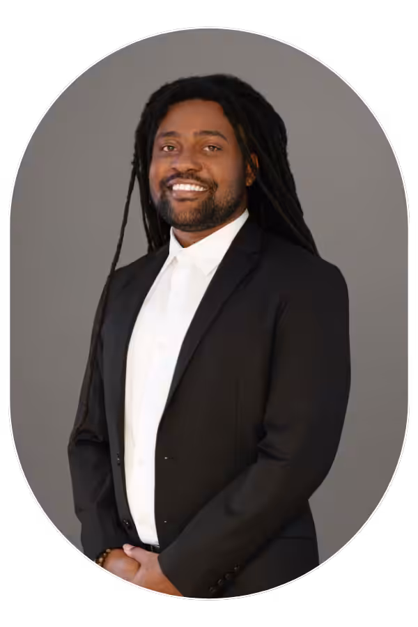 Smiling man with long dreadlocks in a black suit and white shirt, standing against a gray backdrop.
