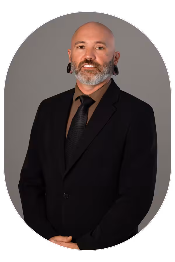 Portrait of a bald man with a gray beard and ear gauges wearing a black suit, brown shirt, and black tie against a gray background.