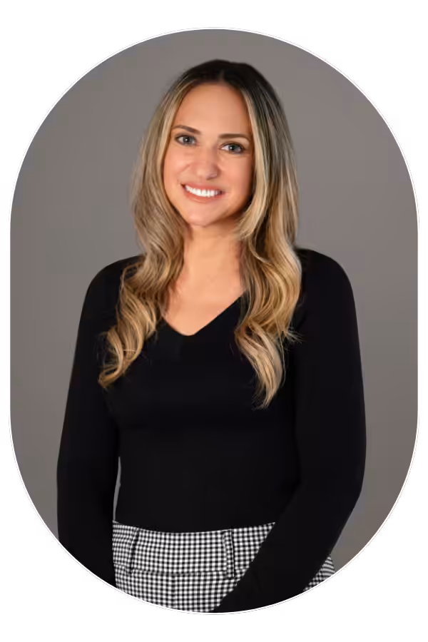 Portrait of a smiling woman with long blonde hair, wearing a black top and checkered skirt, against a gray backdrop.